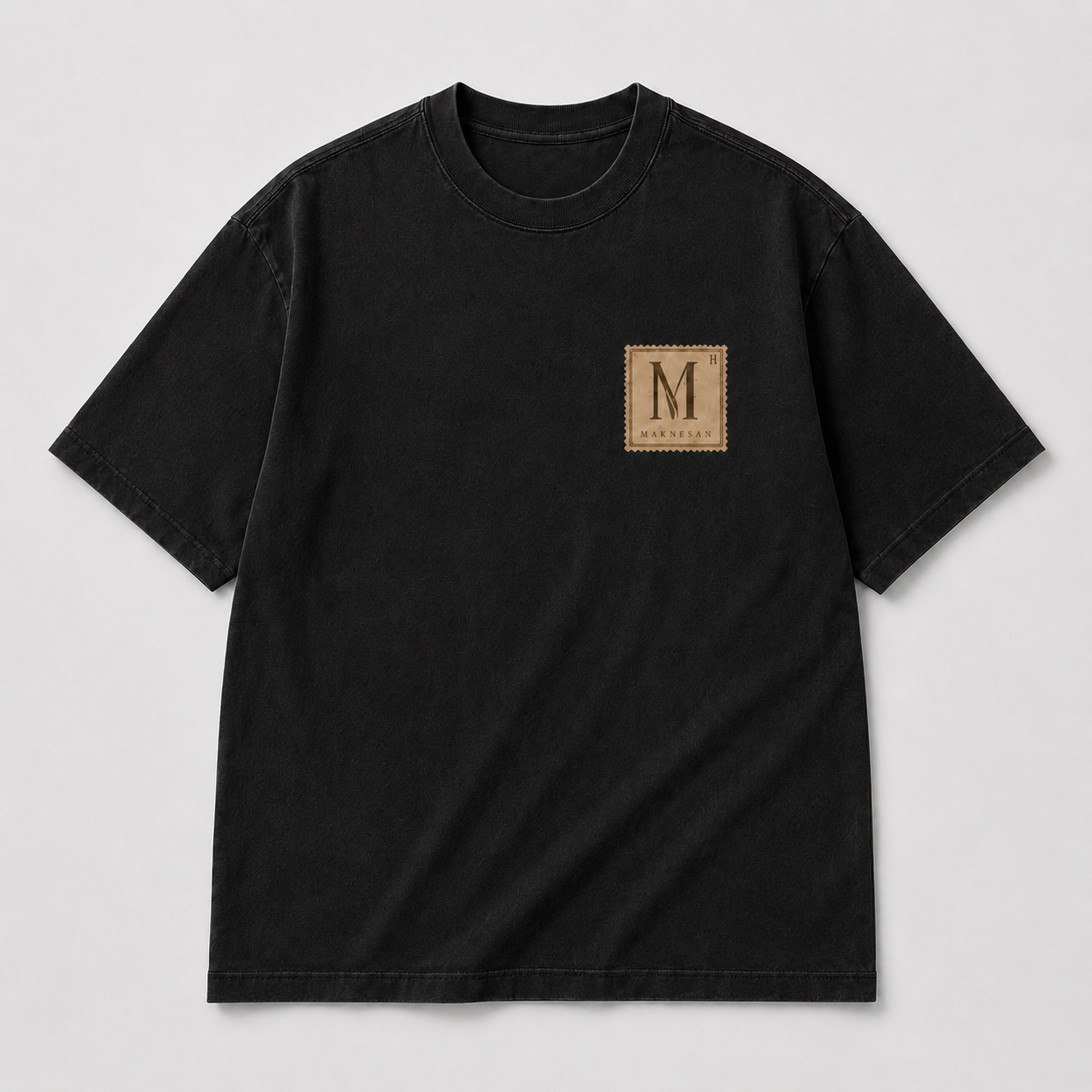 A washed black Maknesan tee laid flat, with the M / MAKNESAN stamp patch at the chest.